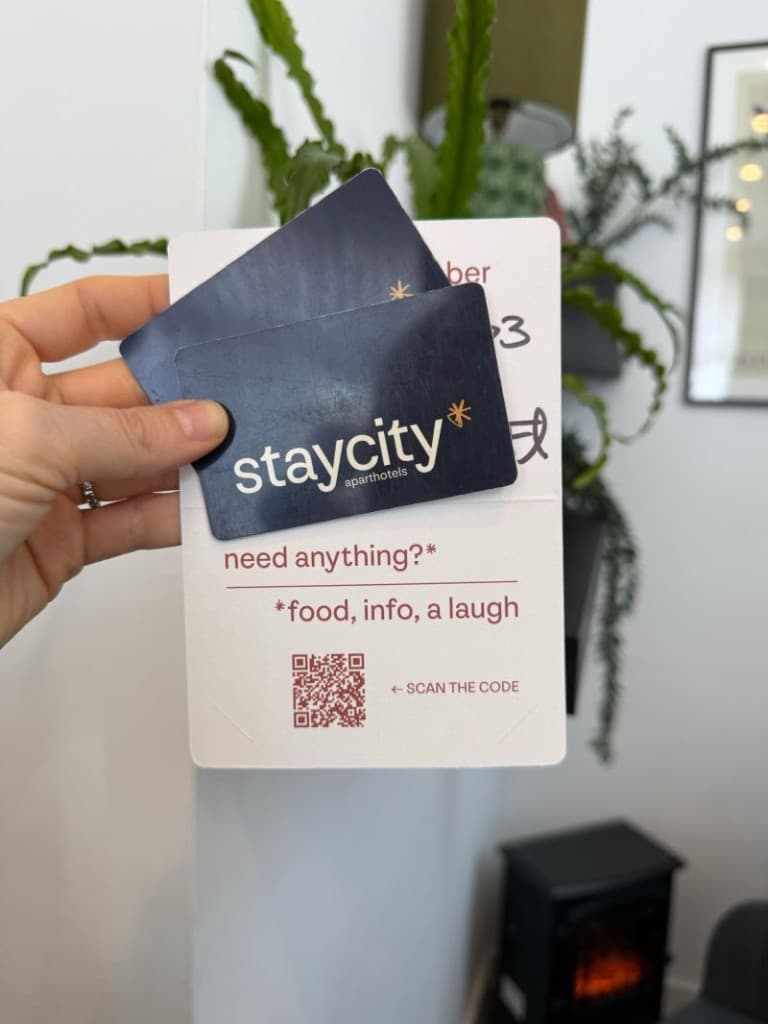 Hand holding Staycity Aparthotels key cards and a white card holder with a QR code labelled need anything? food, info, a laugh, and an arrow that says scan the code